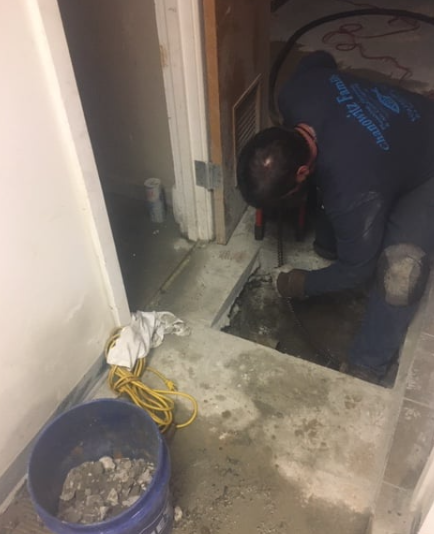 Worker crouched in a small doorway, inspecting concrete floor with a bucket and yellow cord nearby.