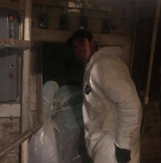 Person in a white protective suit stands in a dim, cluttered room beside a covered object.