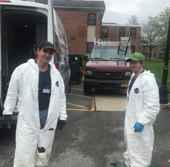 Two people in white protective suits stand beside a van outside a brick building on a wet day