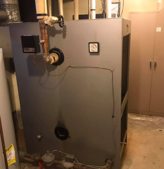 Large gray HVAC furnace in a utility room with pipes, gauges, and a beige cabinet nearby