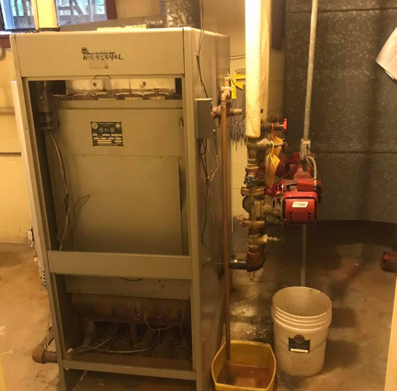 Large industrial HVAC unit in a basement utility room with pipes, valves, and a white bucket on the floor.