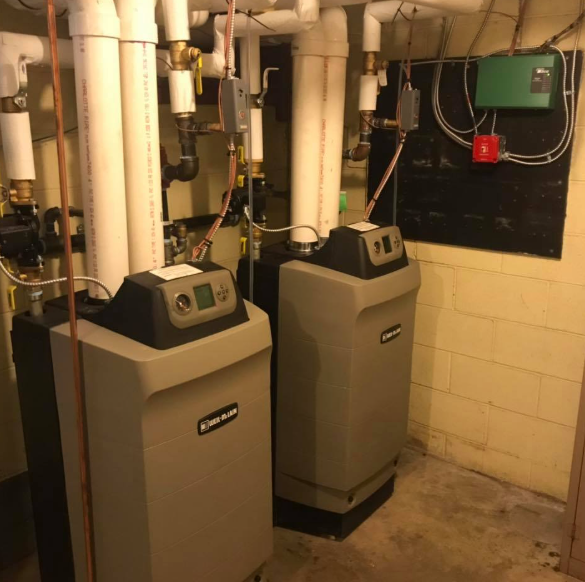 Two gray boiler units in a basement utility room with exposed pipes and valves.