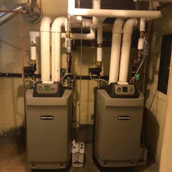 Two gray boiler units with white pipes in a basement mechanical room