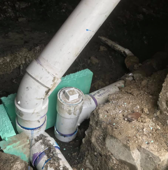 White PVC pipes and a capped cleanout in an open dirt trench beside a concrete foundation.