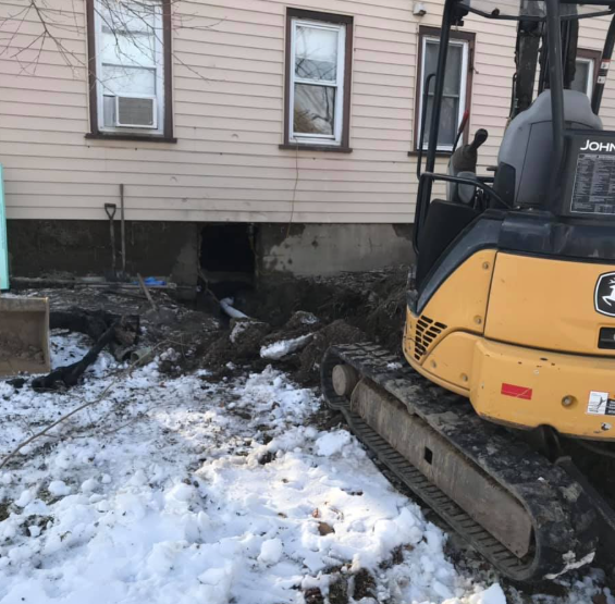 Yellow excavator beside house with muddy trench under the foundation and snow on the ground