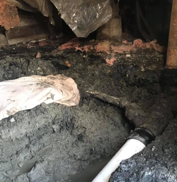 Damaged crawlspace with dark mud, debris, and a white pipe under a wooden floor.