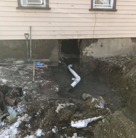 House foundation excavation with a dark crawlspace opening and exposed white pipes in muddy dirt