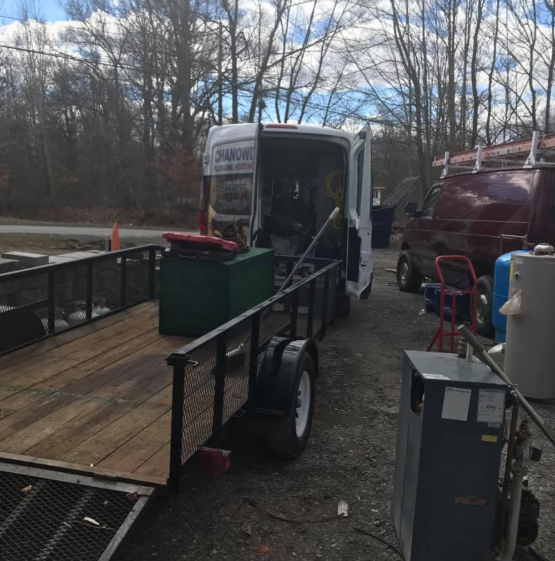 Trailer with green bin and equipment beside an open white van in a gravel lot near trees.