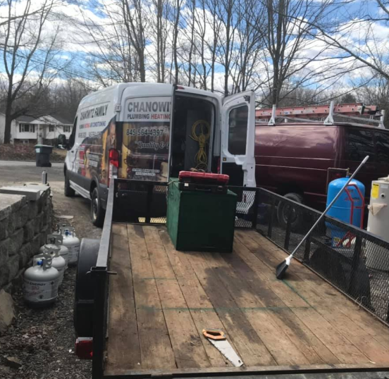White van and maroon tanker on a wooden trailer deck in a yard, with propane tanks and leafless trees nearby