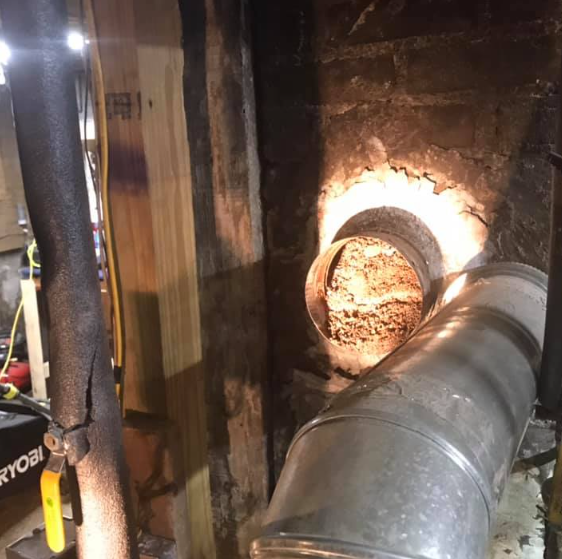Fire-damaged wall with a round duct opening and a large metal pipe in a dark interior space