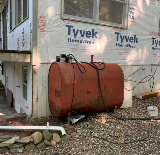 Rusty horizontal tank beside a house under construction, with Tyvek house wrap and debris on the ground.