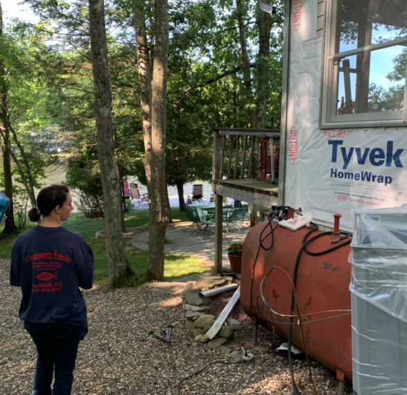 Person standing beside a house under construction with Tyvek wrap, trees and gravel yard in the background
