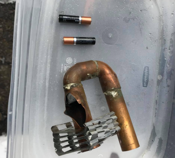 Copper pipe U-shaped tool beside a small black-and-orange battery and a metal spring on plastic tray