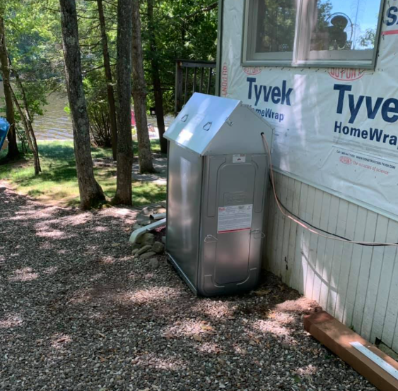 A stainless steel outdoor appliance beside a Tyvek-wrapped house in a wooded yard.