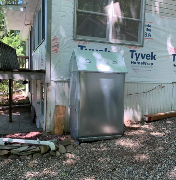 Gray utility cabinet beside a house under construction, with Tyvek wrap and gravel ground.