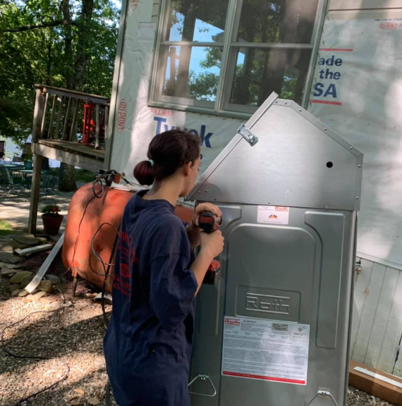 Person working on an outdoor electrical panel beside a building, wearing a dark shirt and orange backpack.