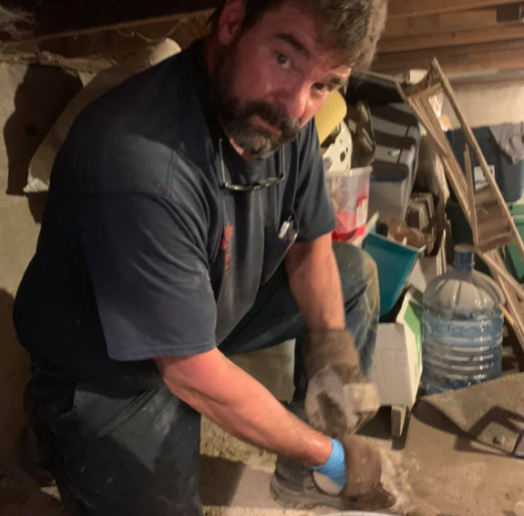 Man kneeling in a cluttered workshop, wearing gloves and preparing a floor surface.