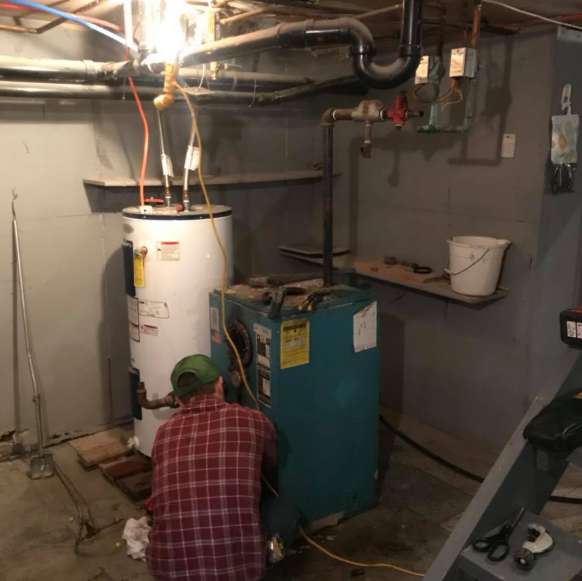 Person kneeling by a white water heater and blue furnace in a basement utility room