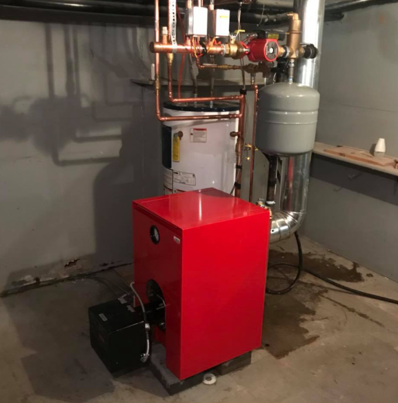 Red boiler and plumbing unit in a basement utility room with exposed pipes and a gray water heater