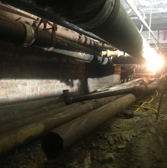 Dim industrial tunnel with large pipes along the walls and floor, bright light at the far end.