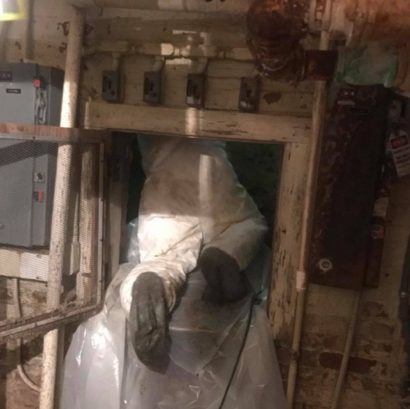 Person in white protective suit working in a narrow industrial hatch with pipes and equipment.