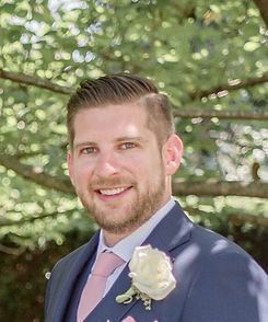 Smiling man in a navy suit and pink tie with a white boutonniere outdoors under trees