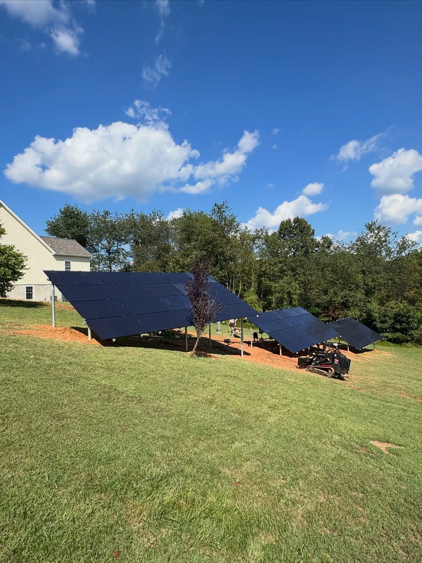 Solar panels installed on a grassy lawn beside a house under a blue sky with clouds