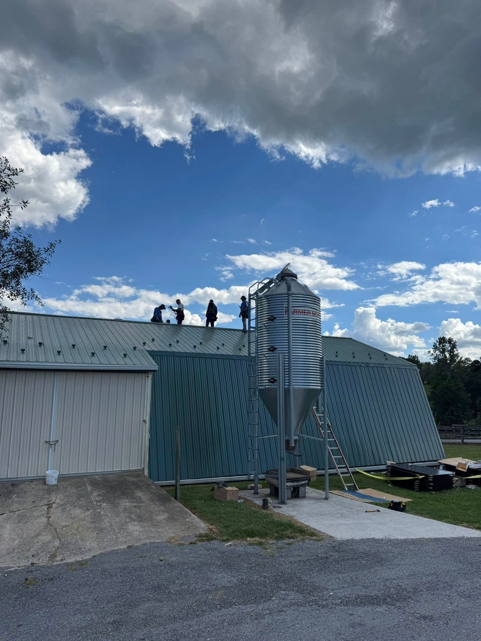 Workers on a metal roof beside a silver silo under a cloudy blue sky