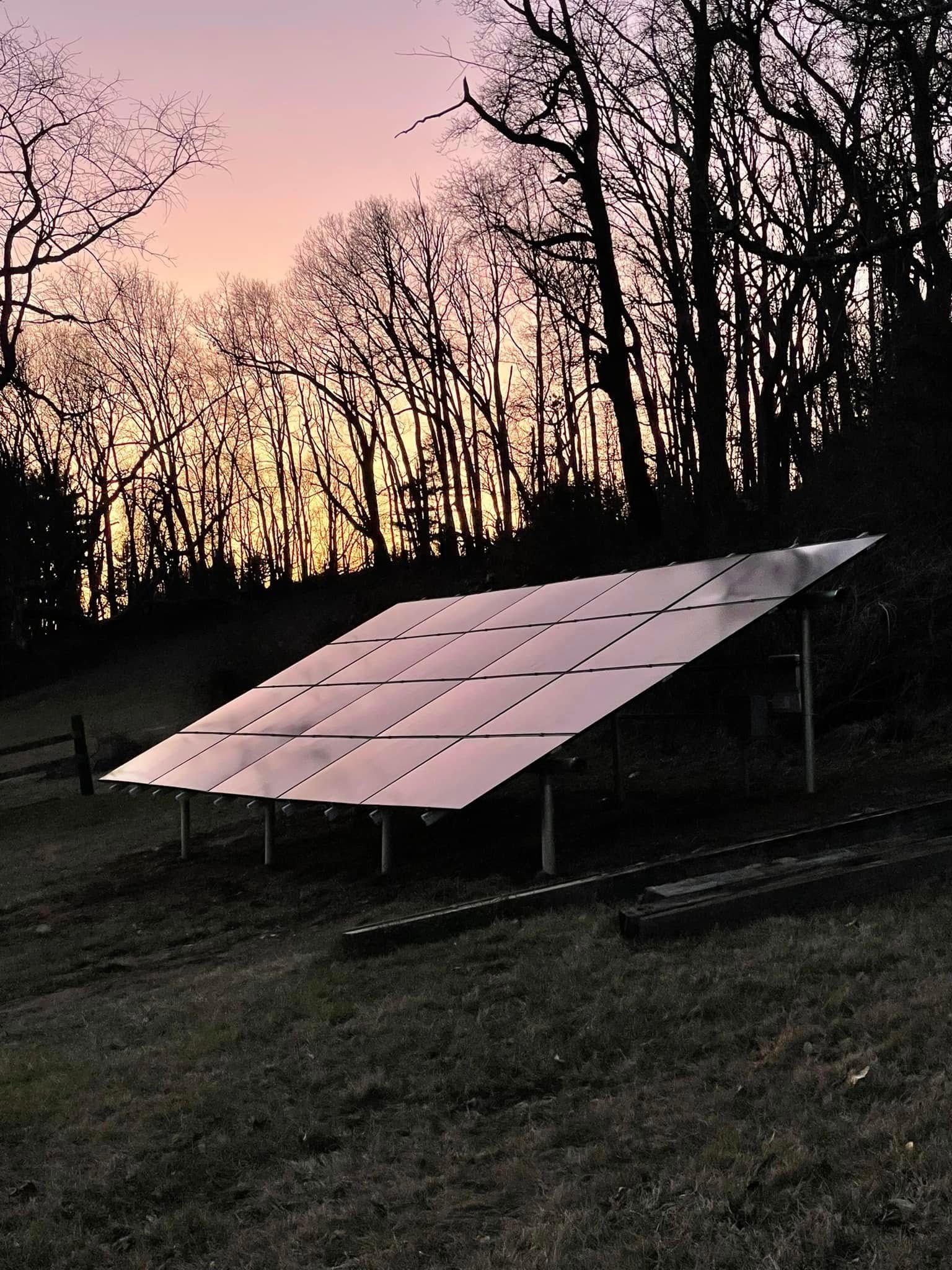 Solar panels mounted outdoors at dusk beneath silhouetted trees against a pink-orange sky
