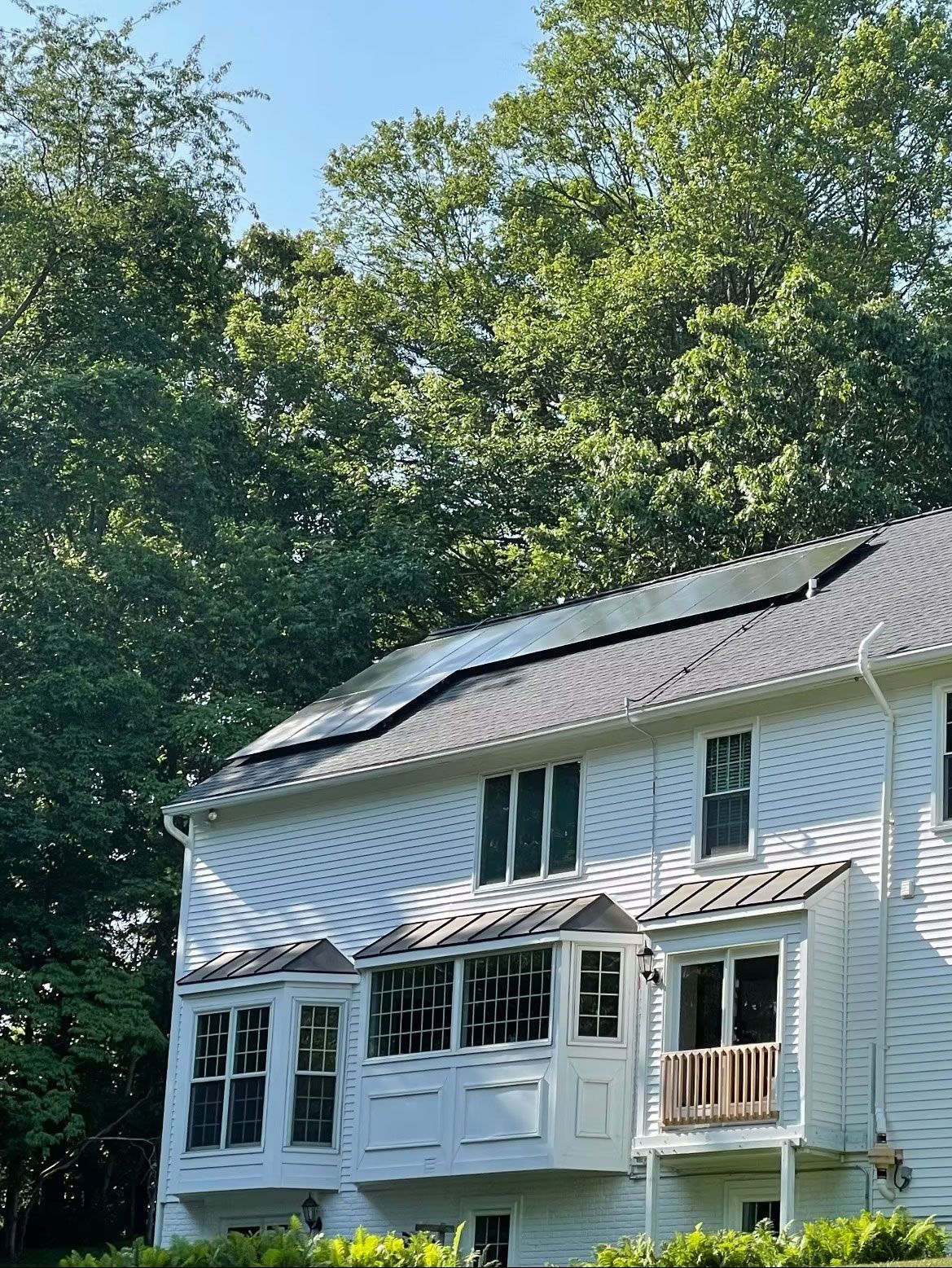 White house with solar panels on the roof, surrounded by tall green trees.