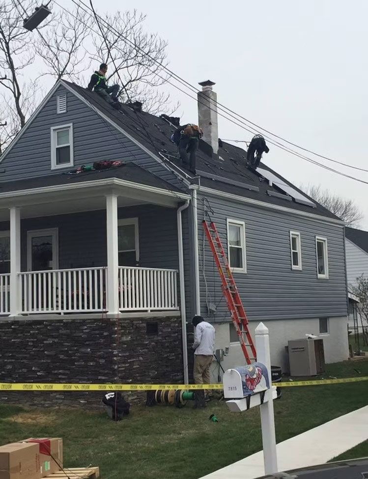 Workers on a blue house roof beside a ladder, with caution tape and a porch below.