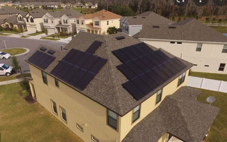 Aerial view of a beige house with dark roof and solar panels in a suburban neighborhood.
