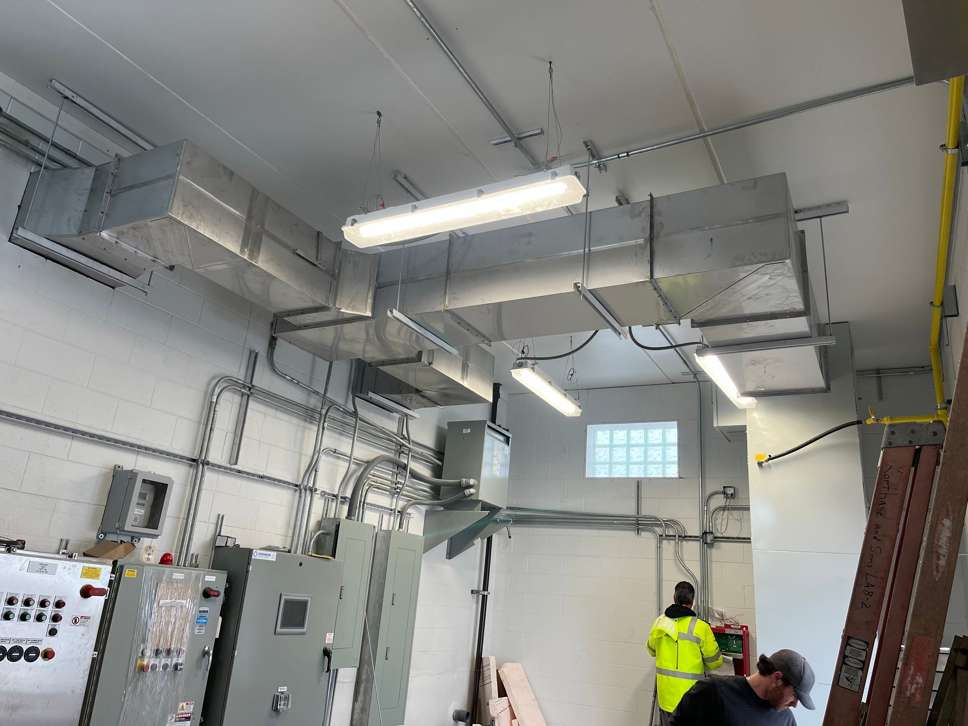 Two workers in an industrial space with large metal HVAC ducts installed on the ceiling and electrical control panels.