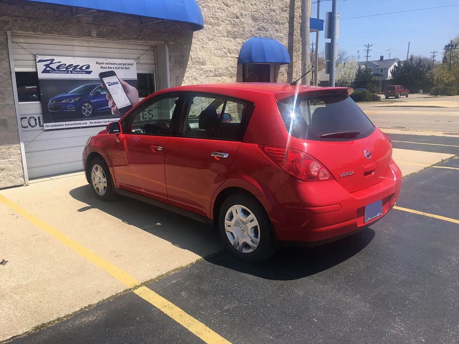 Red Nissan Versa hatchback parked in front of a building with a blue awning.