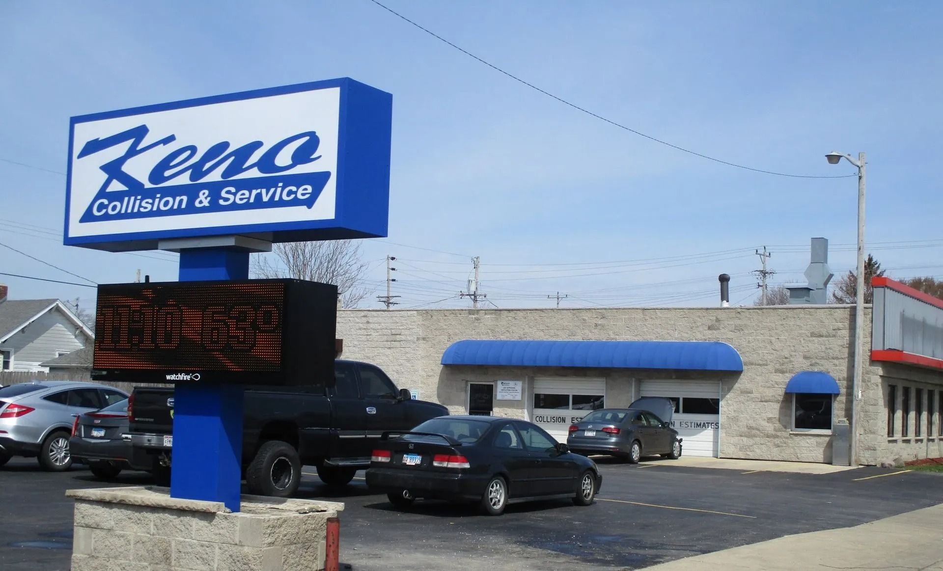 Keno Collision & Service sign on a pole, with a brick building and cars in the parking lot.