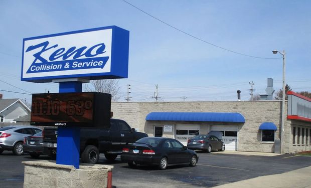 Keno Collision & Service sign on a pole, with a brick building and cars in the parking lot.