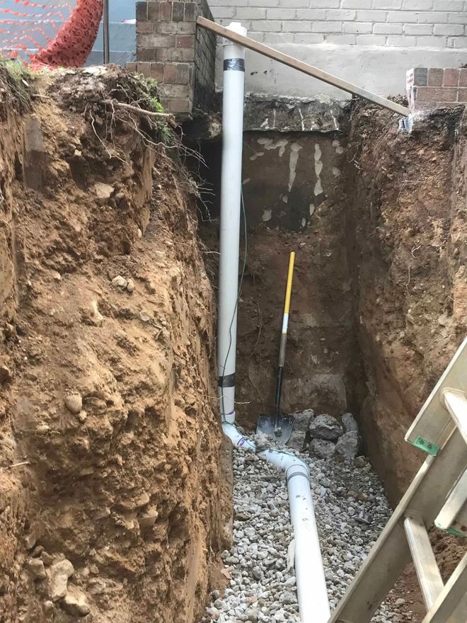 Trench with white PVC pipes and gravel. A pipe runs vertically along a brick wall and connects to a horizontal pipe.