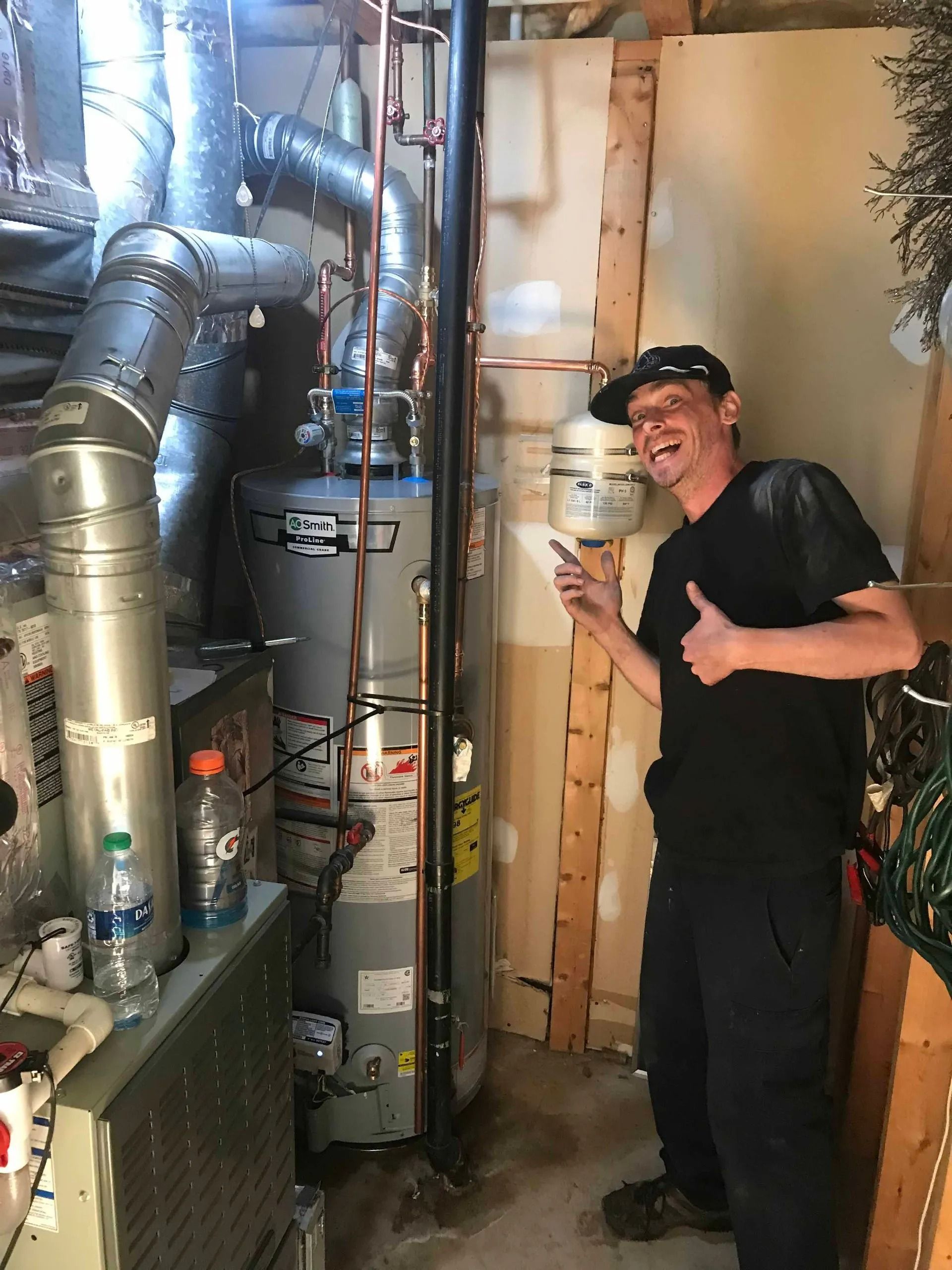 A man in work clothes points at a water heater in a utility room, giving a thumbs up.