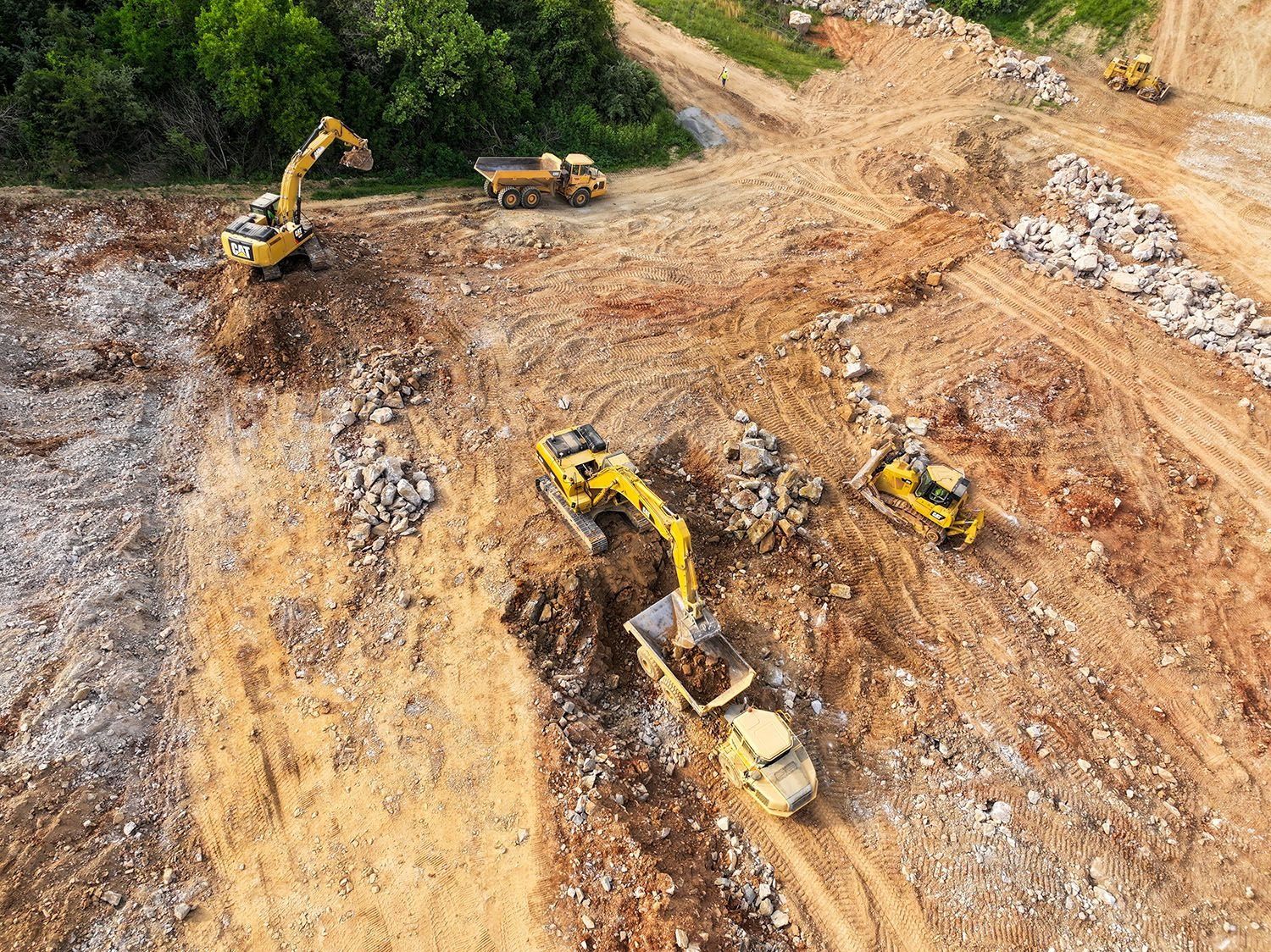 Construction site with yellow excavators and dump trucks on a dirt landscape, clearing land near trees.