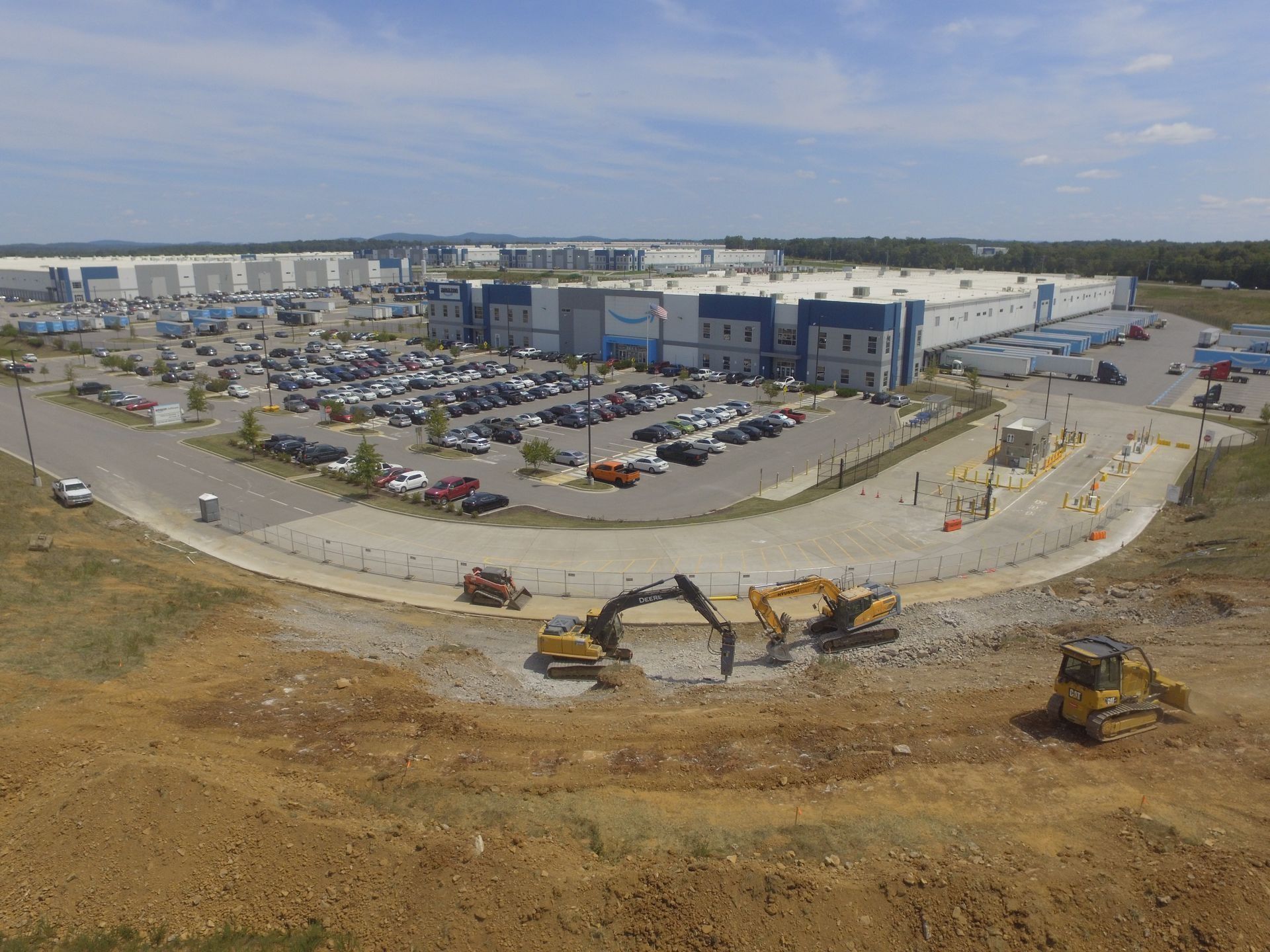 Construction site with heavy machinery in foreground, large industrial building and parked cars in background.