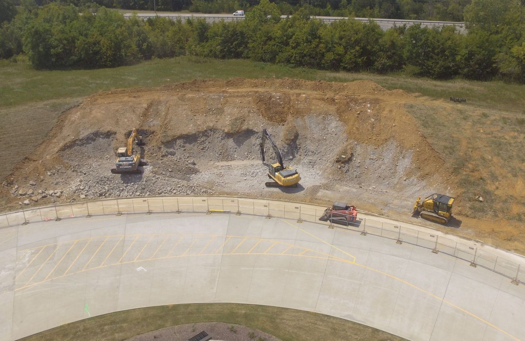 Construction site with excavators on a hillside, working near a curved concrete structure, and a highway in the background.