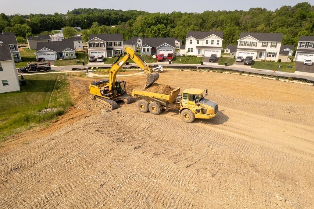Excavator loading dirt into a dump truck on a construction site, residential neighborhood in the background.