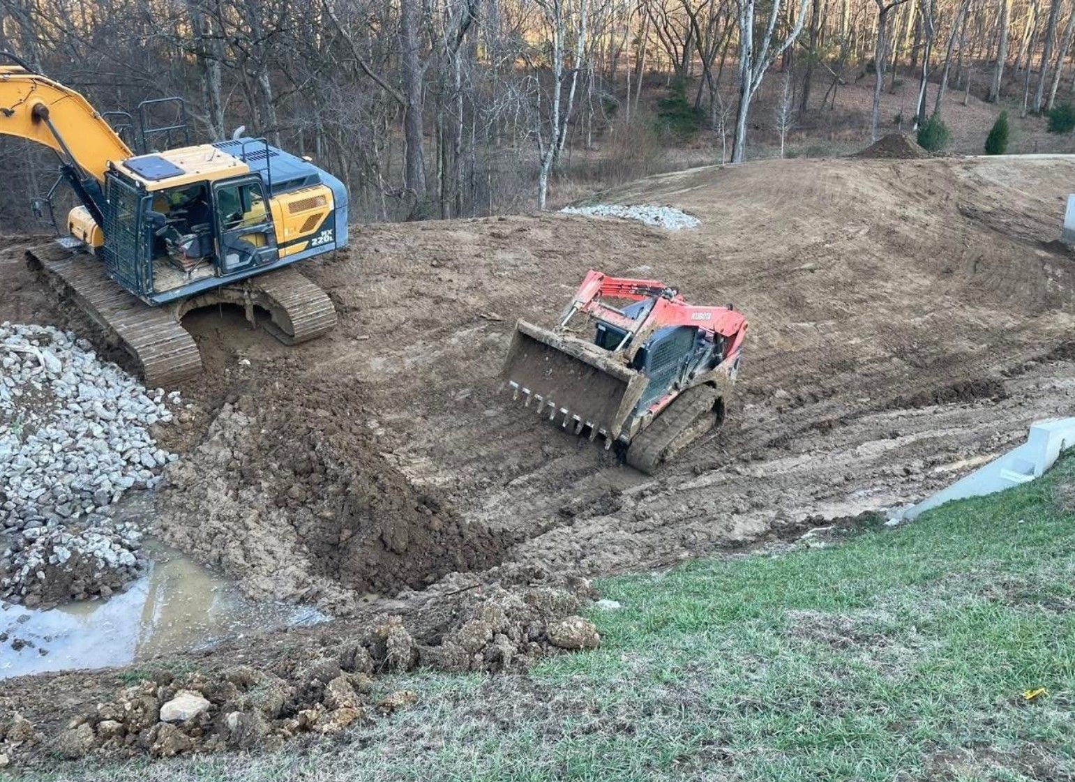 Excavators on a muddy construction site, moving earth near a grassy area and trees.