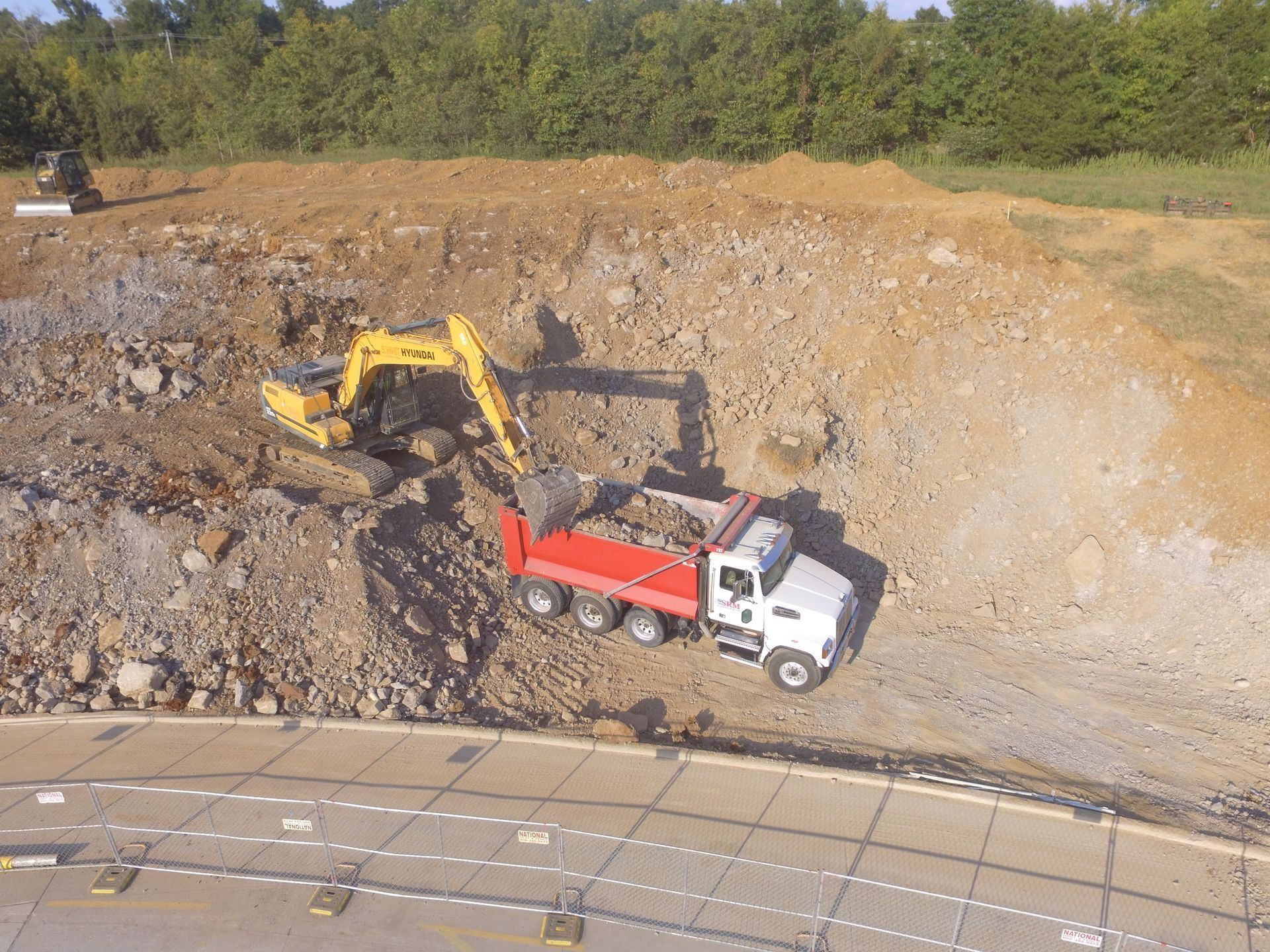 Yellow excavator loading a red dump truck on a construction site with a rocky hillside.