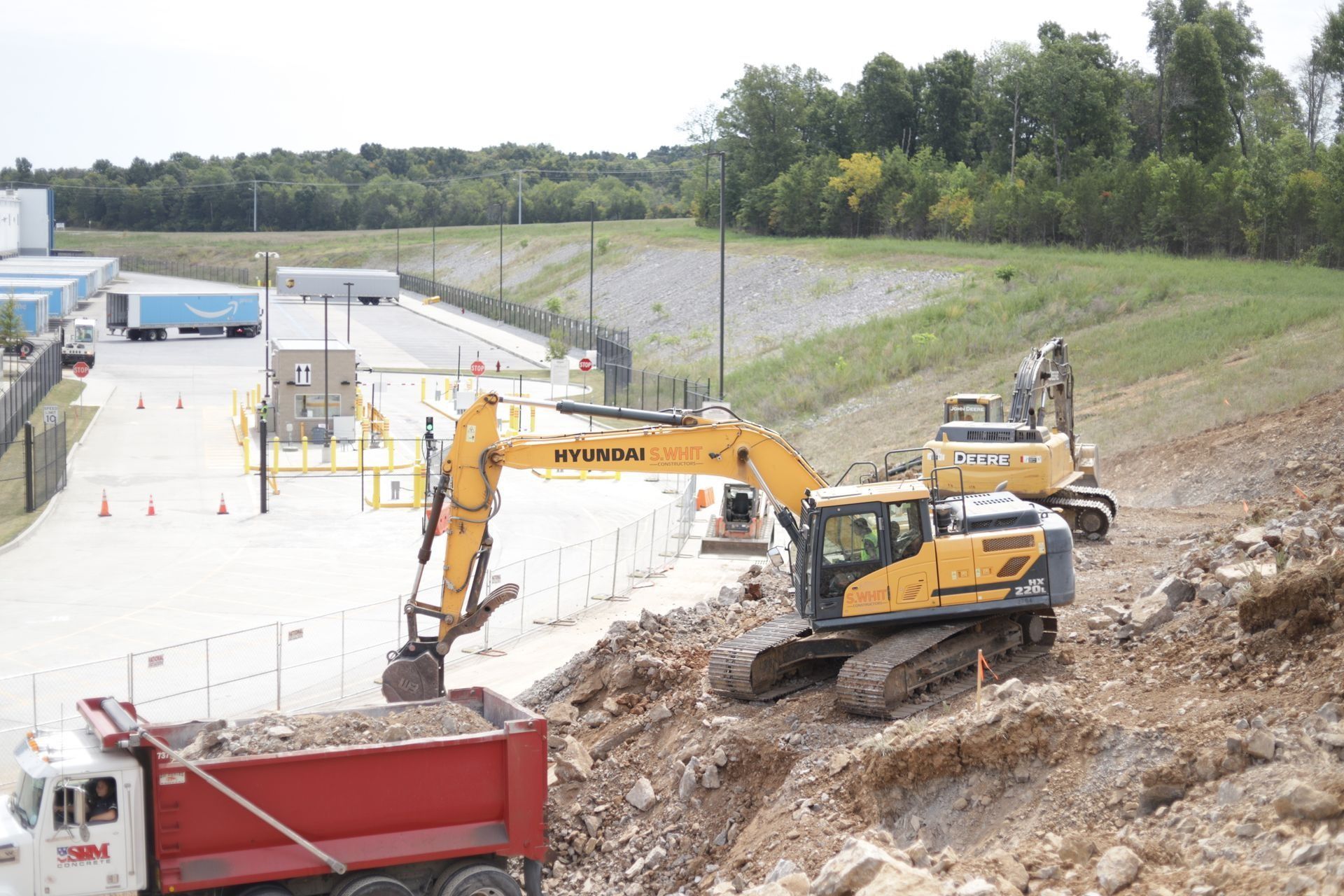 Excavator loading dirt into a dump truck at a construction site, with an Amazon facility in the background.