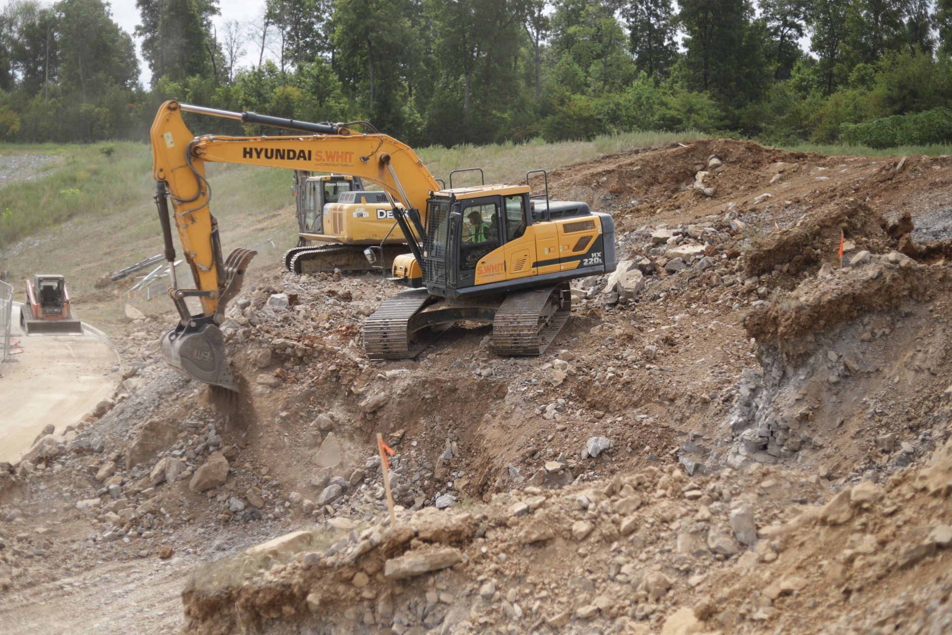 Yellow excavator working on a dirt mound in a construction site, with a second excavator and a small bulldozer in the background.