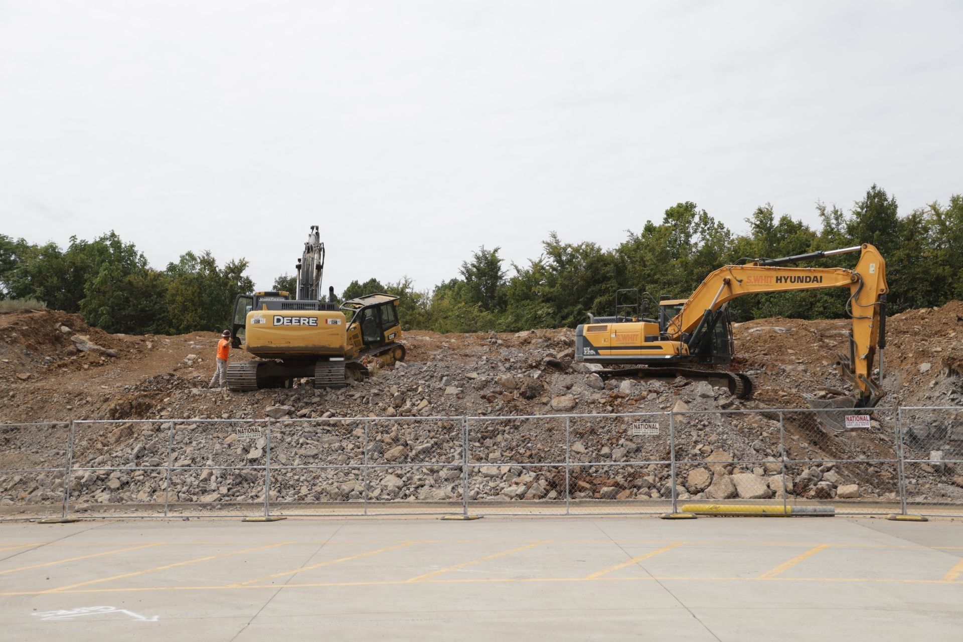 Two excavators on a rocky construction site with trees in the background under a cloudy sky.