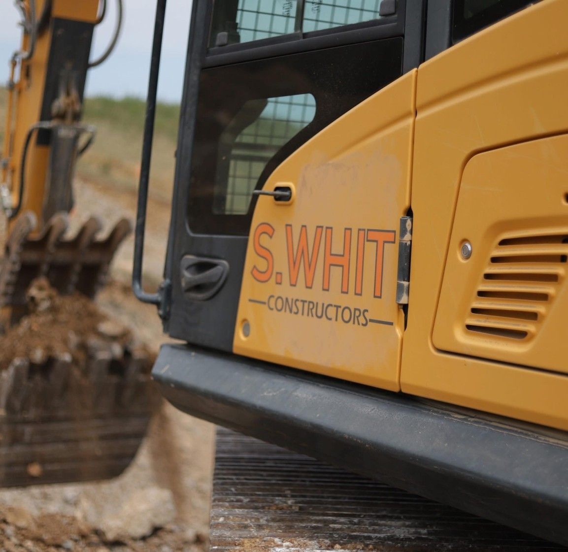 Yellow excavator digging earth; S. WHIT Constructors logo on the side.
