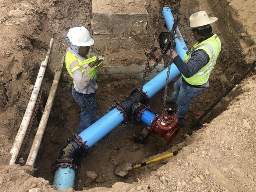 Two workers installing large blue water pipes in a trench, using tools and wearing hard hats and vests.