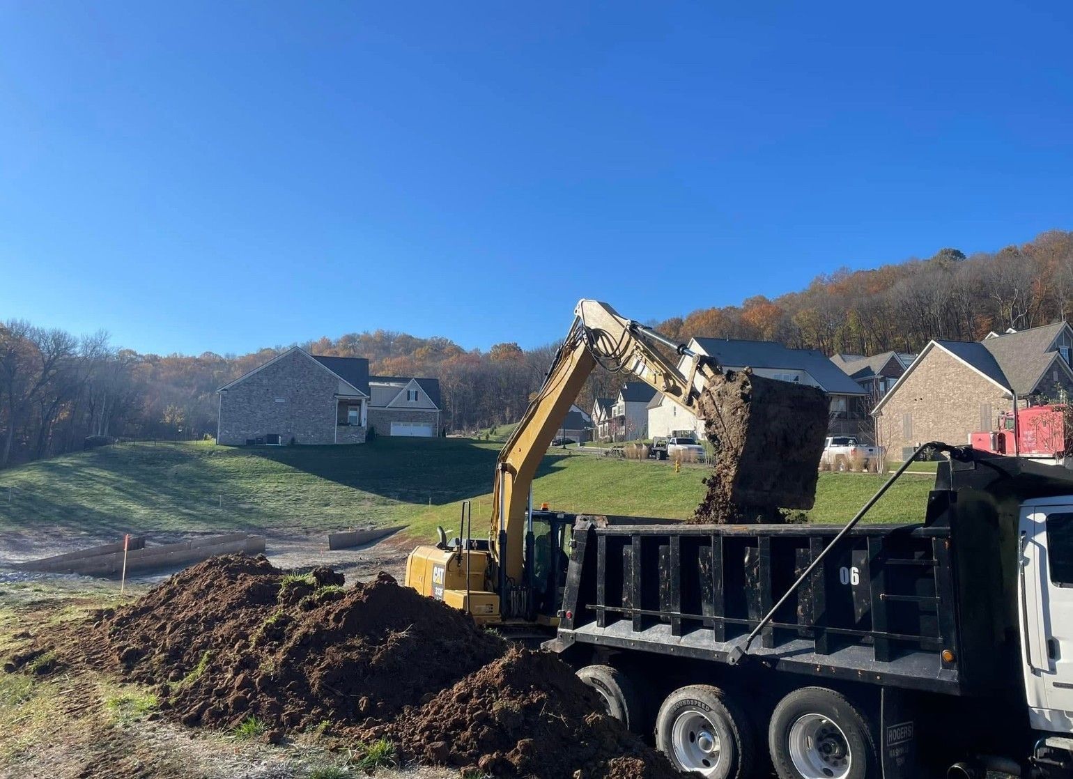 A yellow excavator loads dirt into a dump truck on a construction site with houses in the background.